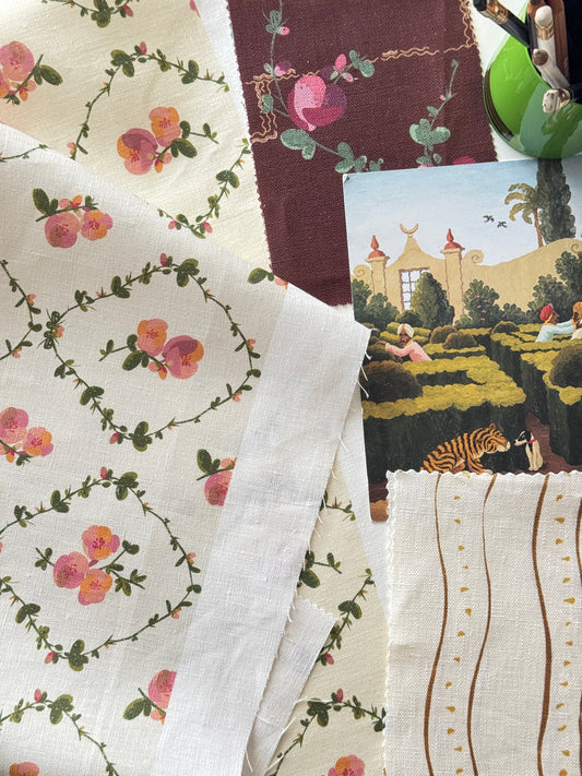 A styled flat-lay of multiple fabric swatches and textile pieces, including floral prints, striped linen, and a small illustrated card, arranged on a neutral surface.