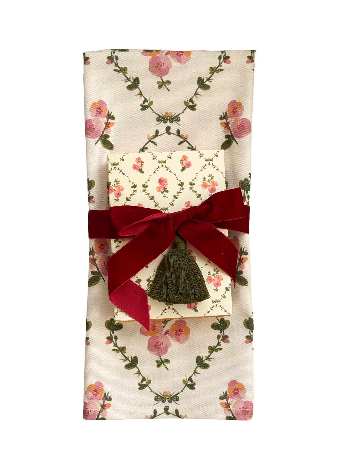 A folded cream-colored tea towel featuring a pink rose and green vine floral pattern, wrapped with a dark red velvet ribbon and a dark green tassel bow, with a smaller matching patterned matchbox tucked inside.
