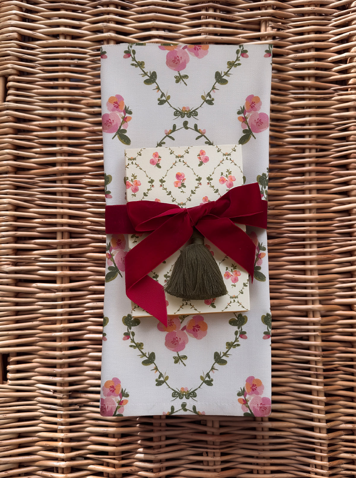 A folded gift set of decorative matchbox and tea towel  with a pink rose and green vine floral pattern, tied with a deep red velvet ribbon and bow, and finished with a dark olive tassel, set on a woven wicker surface.