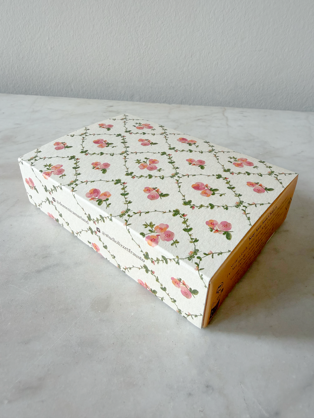 A rectangular cardboard box with a white background and a repeating pink floral vine pattern, sitting on a light marble surface against a neutral wall.