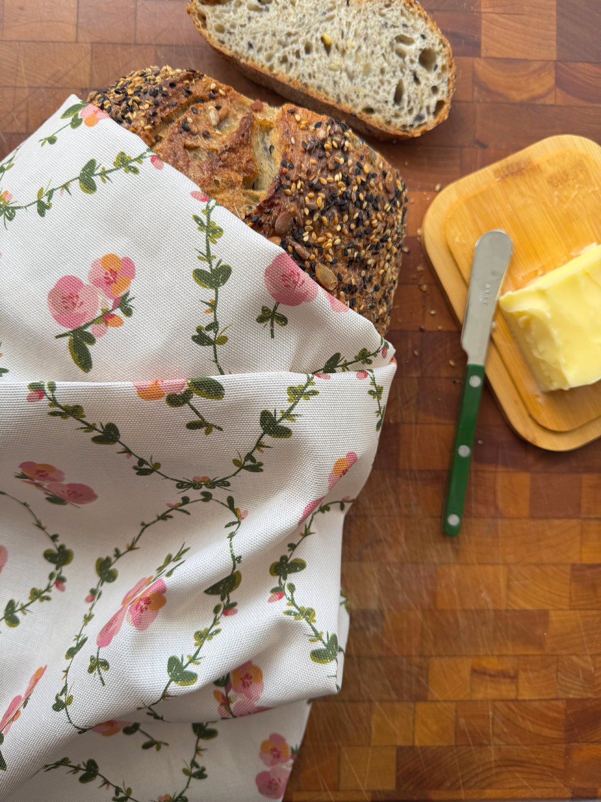 A floral bread cloth tea towel with pink flowers and green vines draped over a loaf of bread on a wooden cutting board, with sliced bread, butter, and a butter knife nearby.