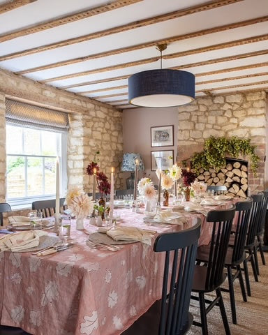 A styled dining room with a long table covered in a dusty pink floral tablecloth, set with plates, candles, and flowers. Black spindle-back chairs surround the table, set against a rustic background of stone walls, exposed wooden ceiling beams, and a hanging navy blue drum chandelier.