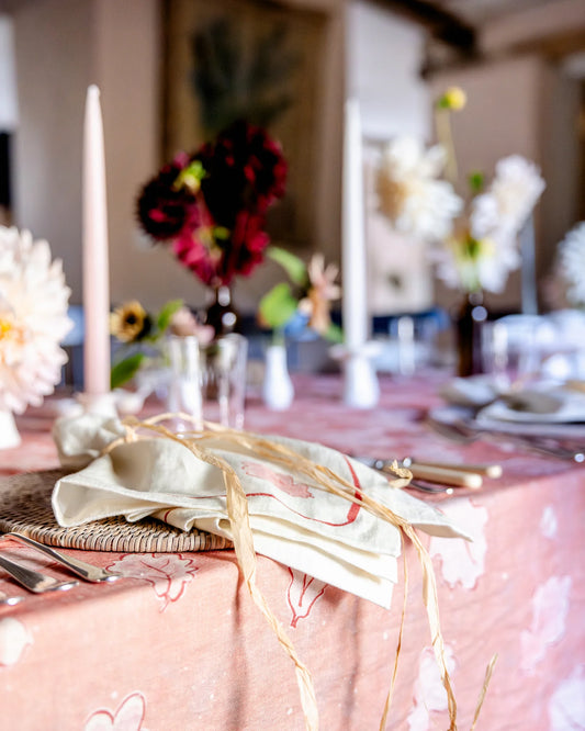A styled dining table setting with a blush pink floral tablecloth, cream napkins tied with twine, white taper candles, and floral centerpieces in a softly blurred indoor environment.
