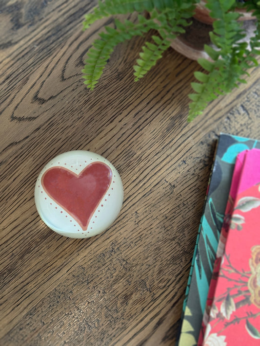A round glass decoupage paperweight featuring a pinkish-red heart in the center, placed on a wooden tabletop next to a potted fern and floral notebooks.